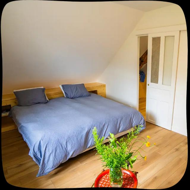 Chambre 1 - Lit double avec des draps bleu clairs. On voit la porte d'entrée au milieu à droite. On voit un petit tabouret rouge en avant plan, devant le lit, avec un bouquet de fleurs verte.