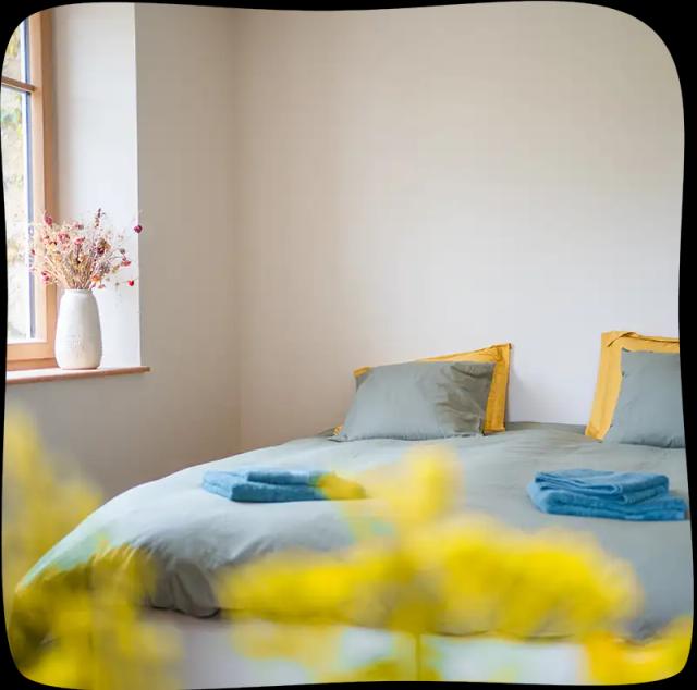Chambre 2 – Vue proche du lit avec des draps bleu tournant vers le gris clair avec 4 coussins dont 2 de la même couleur et deux jaunes. On peut aperçevoir un bout d'une fenêtre lumineuse. Il y a des essuies de bain bleus sur le lit. Le premier plan est un flou de fleurs au bout jaune