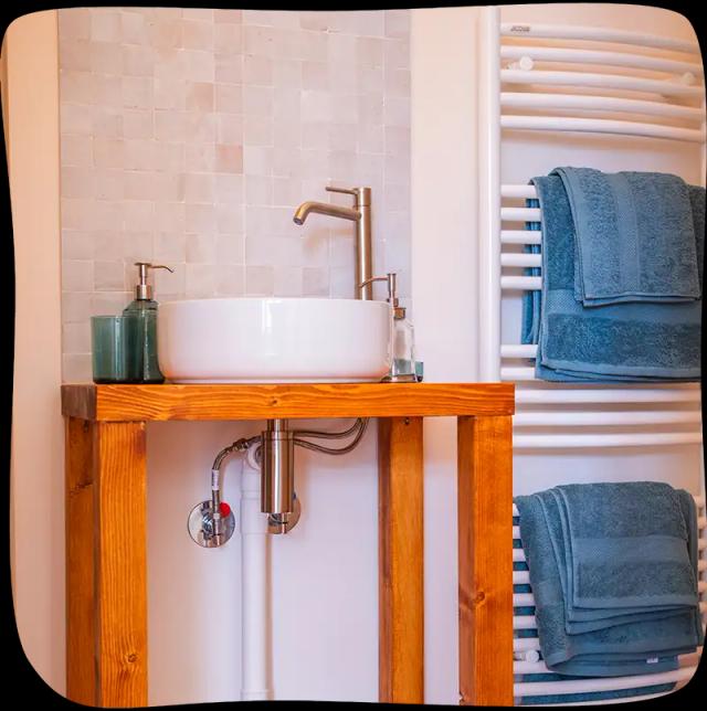 Chambre 2 – Evier de la salle de bain sur un meuble en bois. On y retrouve un savon main et un verre. Il y a un porte serviette mural chauffant à droite avec 2 paires d'essuies bleu