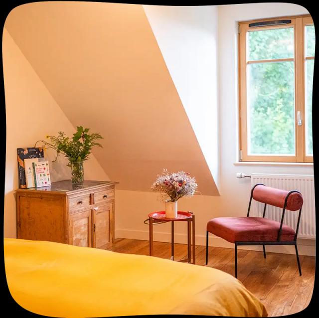 Chambre 3 – On voit le bout du lit avec des draps jaunes moutarde en premier plan et un fauteuil rouge ainsi qu'une armoire en bois et un tabouret rouge avec un pot de fleurs jaunes et vertes eu milieu du plan. Le fauteuil se trouve juste devant une grande fenêtre lumineuse