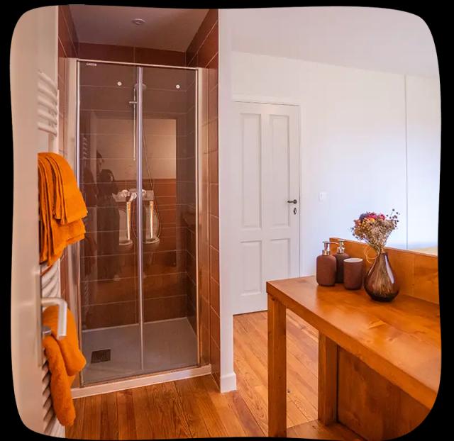 Salle de douche de grande taille ouverte sur la chambre avec un meuble en bois en premier plan sur la droite. On voit aussi un porte serviette mural chauffant à gauche avec des essuies de bain orange.