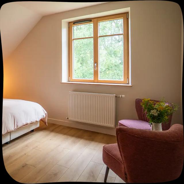 Une grande fenêtre lumineuse avec un radiateur en dessous au miliau, un bout du lit double aux draps rose pâle à gauche et deux fauteuils rouge à droite. On peut aperçvoir un pot de fleur entre les deux fauteuils.